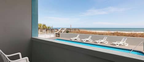 A balcony with a pool, white chairs, and a view of the beach.
