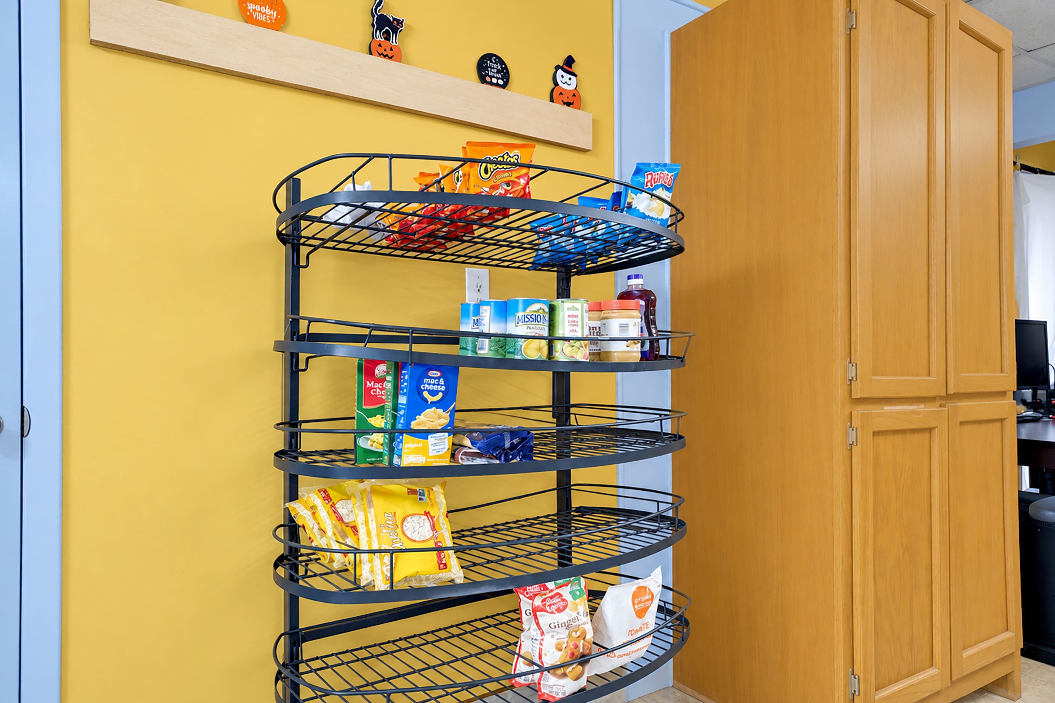 Un estante metálico de varios niveles contra una pared amarilla, con diversos alimentos y artículos del hogar.