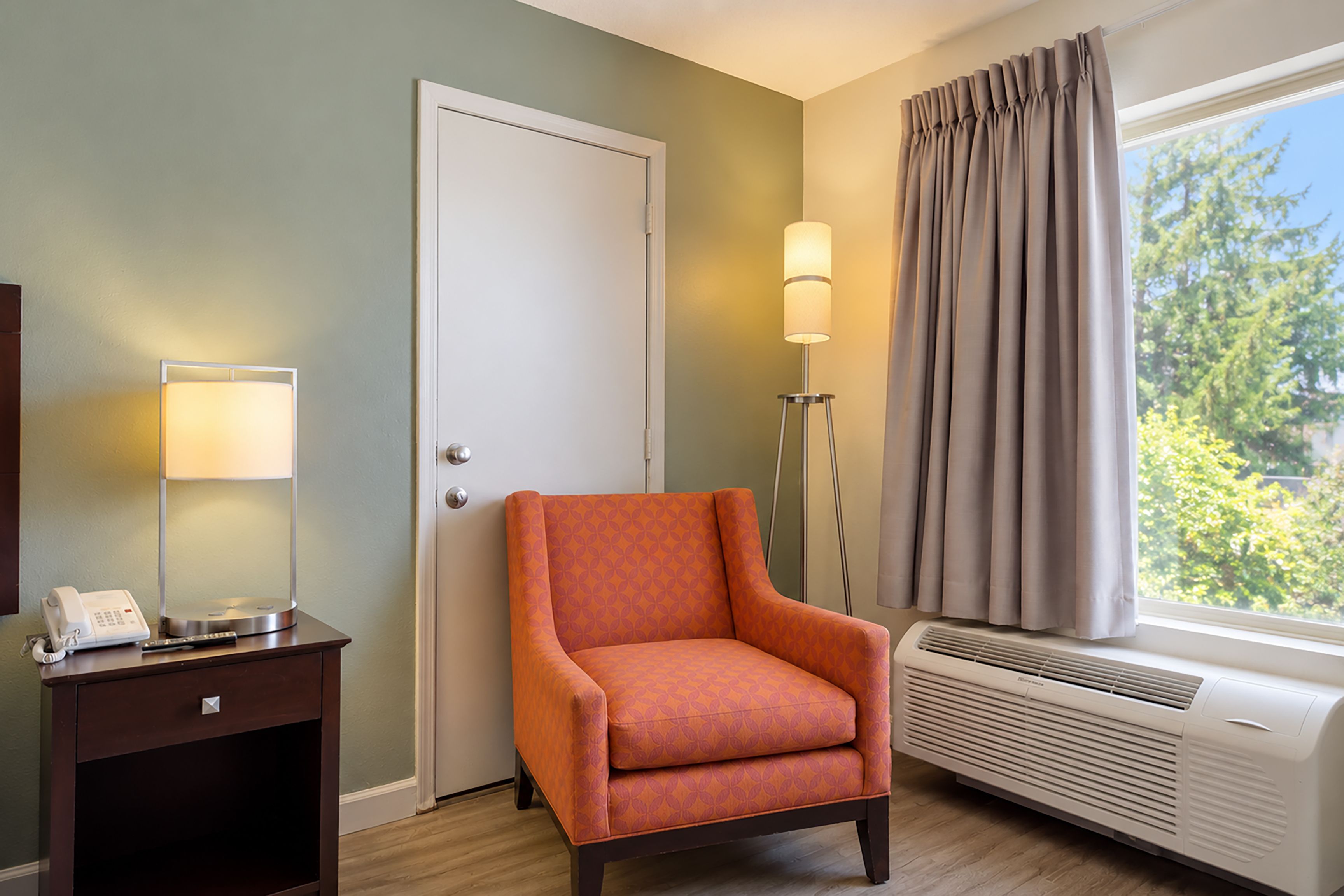 Una habitación con una silla, un escritorio con teléfono, una lámpara, una ventana con cortinas y un equipo de aire acondicionado.