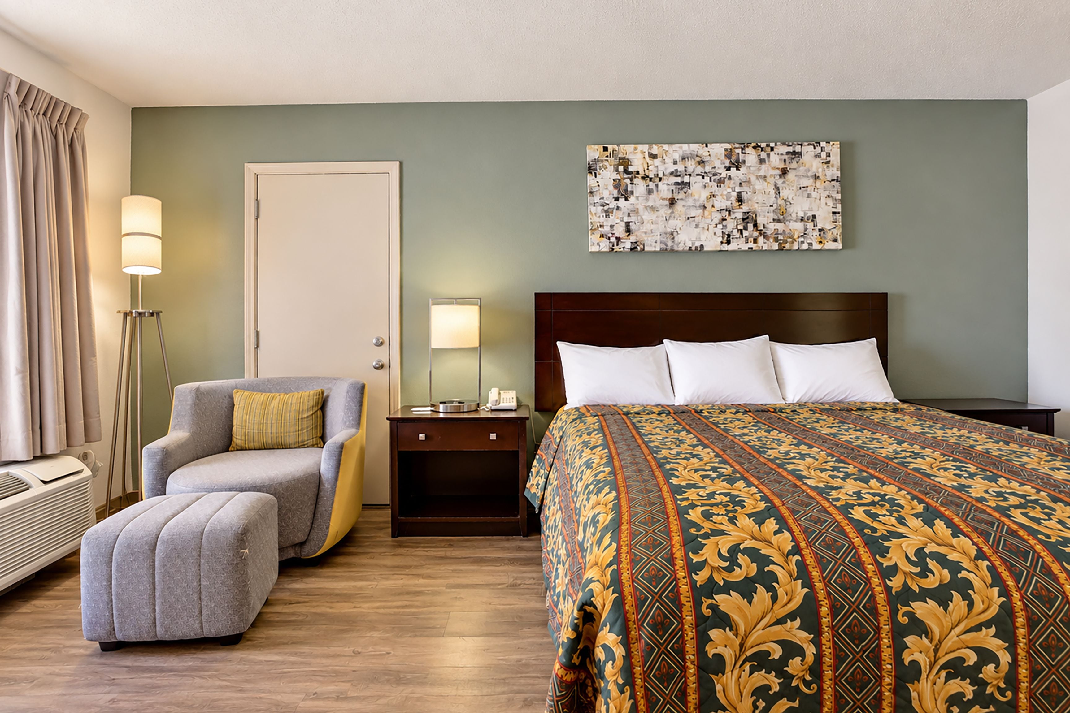 Habitación de hotel con cama, una silla, una mesita de noche, lámpara y un cuadro en la pared.