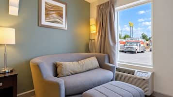 Habitación de hotel con un sofá gris, un cuadro abstracto enmarco, una lámpara, un ventanal con vista a un camión U-Haul y un termostato.
