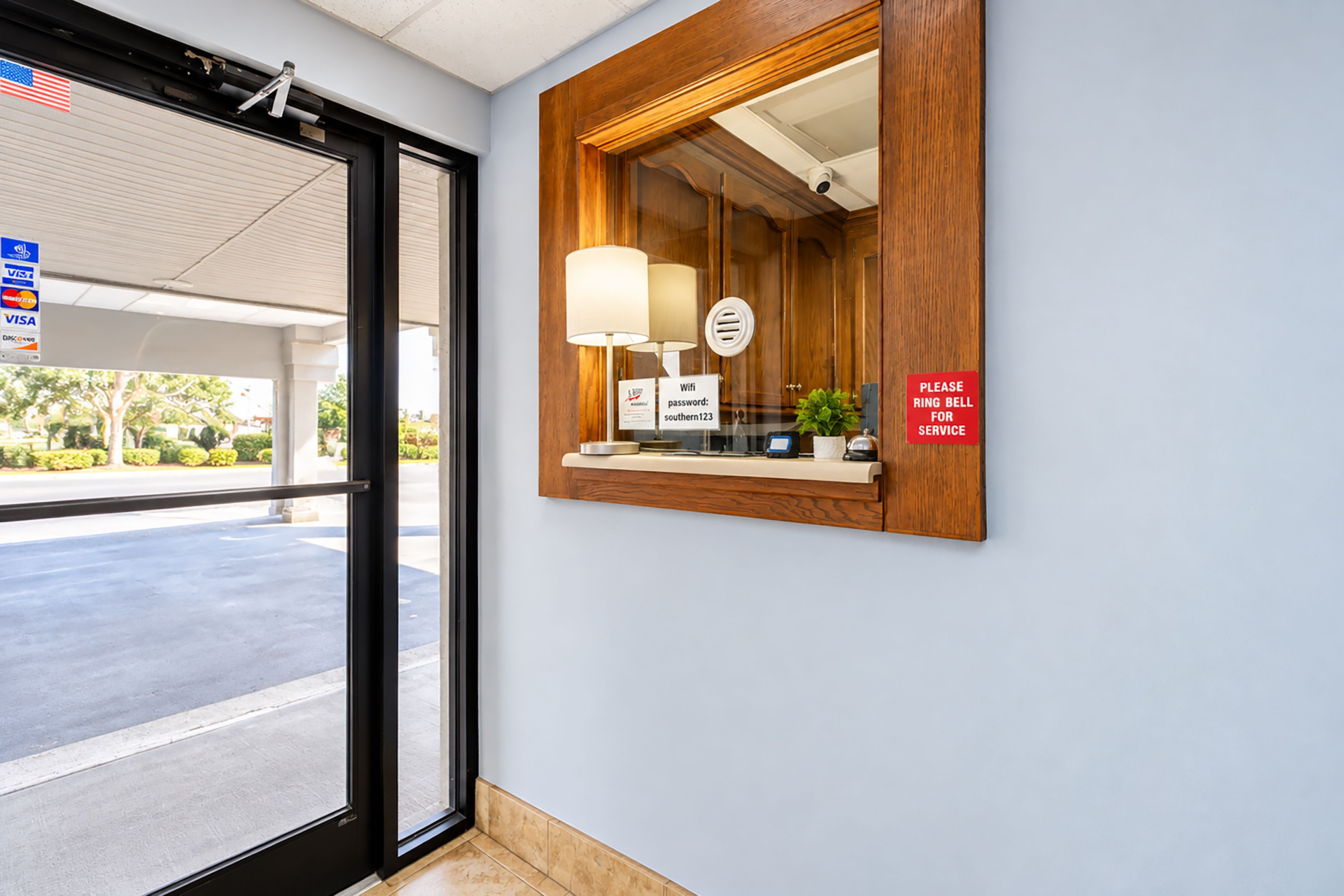 Een hotel receptie met een houten receptiebalie, een spiegel, een bord met de WiFi-wachtwoord en een bord waarop staat om voor service de bel te luiden.
