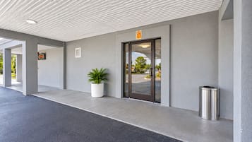 L’entrée d’un immeuble avec un toit plat, un pot de fleurs, une poubelle et une porte au-dessus de laquelle est affichée une enseigne.