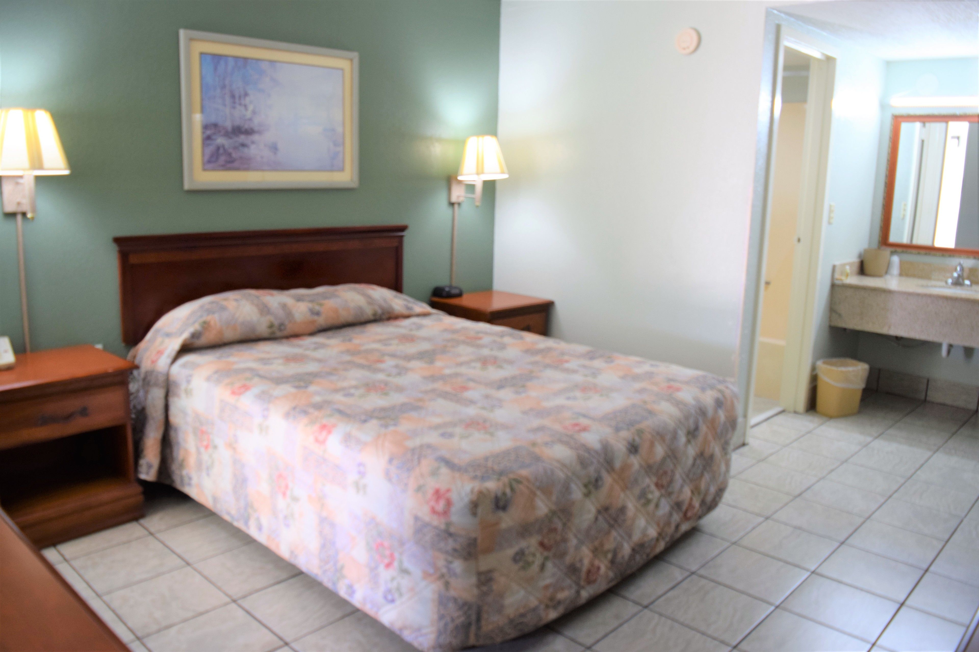 A bedroom with a bed, nightstands, a lamp, a painting on the wall, and a bathroom with a sink and mirror.