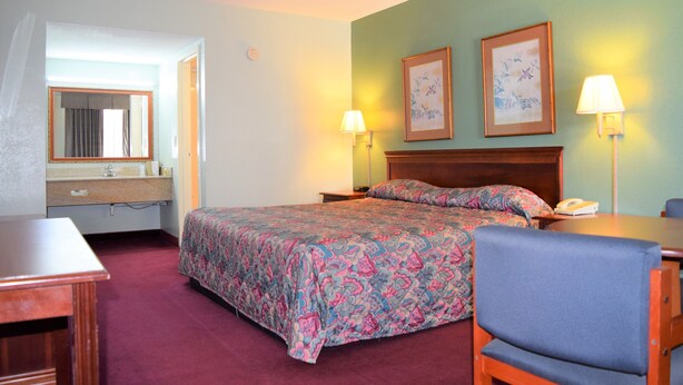 A hotel room with a bed, bedside lamps, a chair, a desk, and a bathroom with a mirror and sink. - Liberty Inn (Waynesboro)