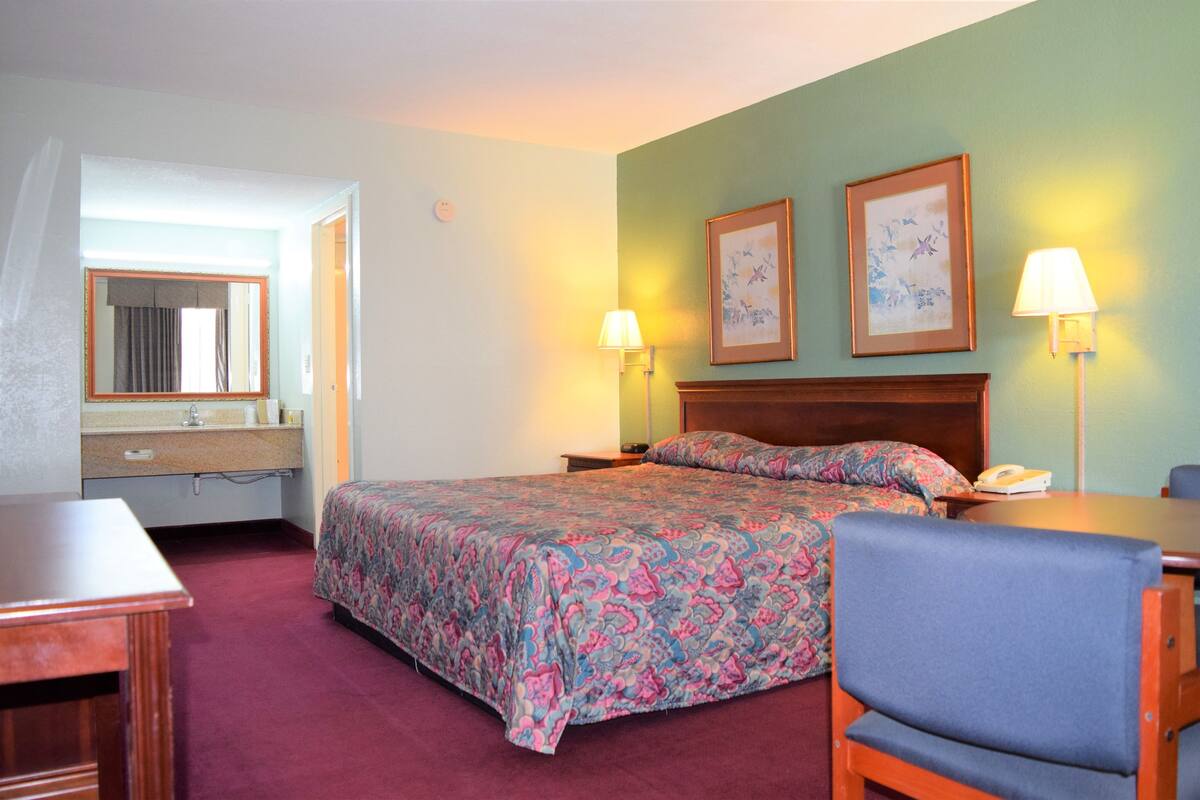A hotel room with a bed, bedside lamps, a chair, a desk, and a bathroom with a mirror and sink.