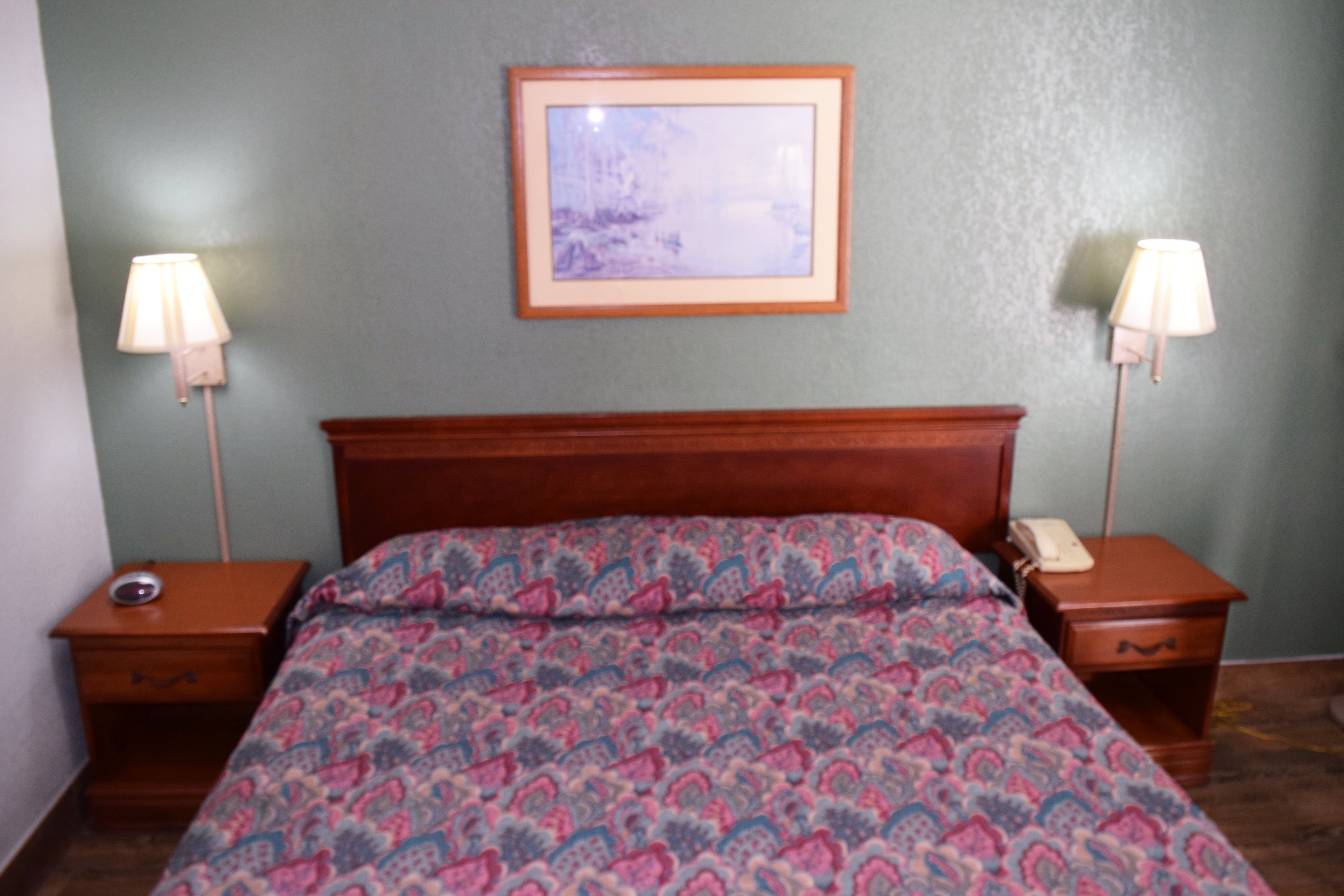 A bedroom with a wooden headboard, a patterned bedspread, two bedside tables with lamps, and a framed picture on the wall.
