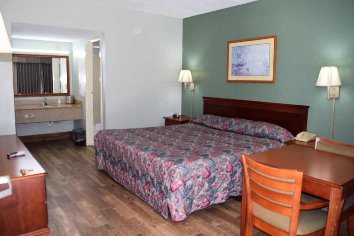A hotel room with a bed, a desk, a chair, a dresser, a mirror, a lamp, and a painting on the wall.