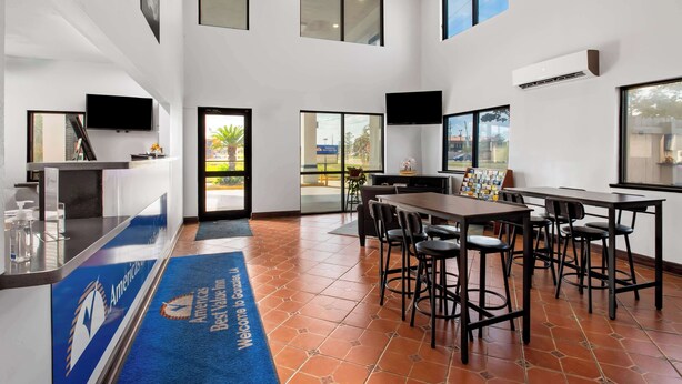 A hotel lobby with a reception desk, a large dining table with chairs, and a blue carpet with the hotel logo. - Americas Best Value Inn Gonzales (Gonzales)