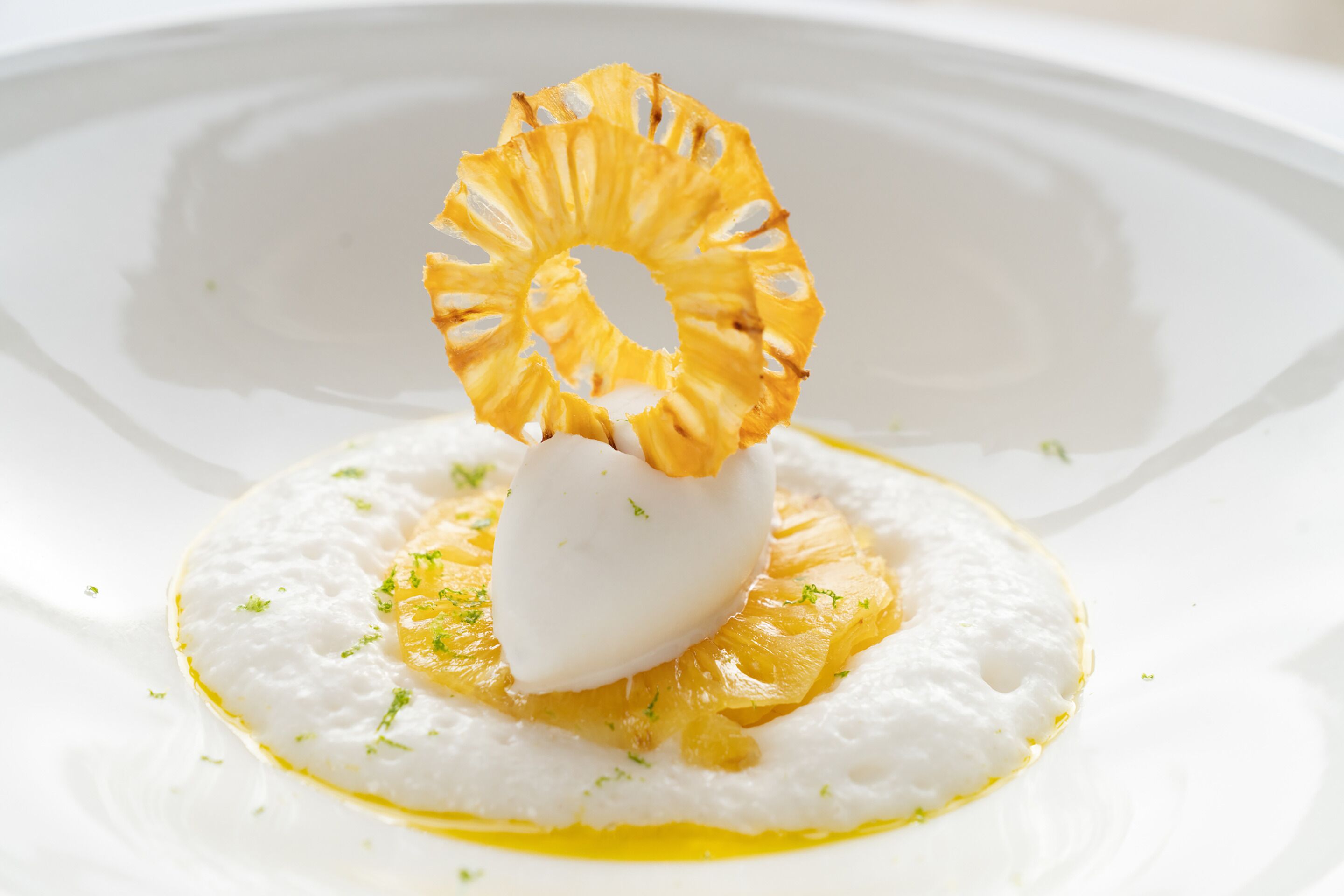 Un plato blanco con un alimento esférico blanco, adornado con hierbas verdes y un aro de piña, sobre una superficie blanca.