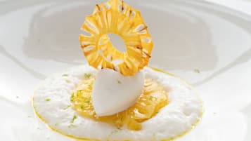 Un plato blanco con un alimento esférico blanco, adornado con hierbas verdes y un aro de piña, sobre una superficie blanca.