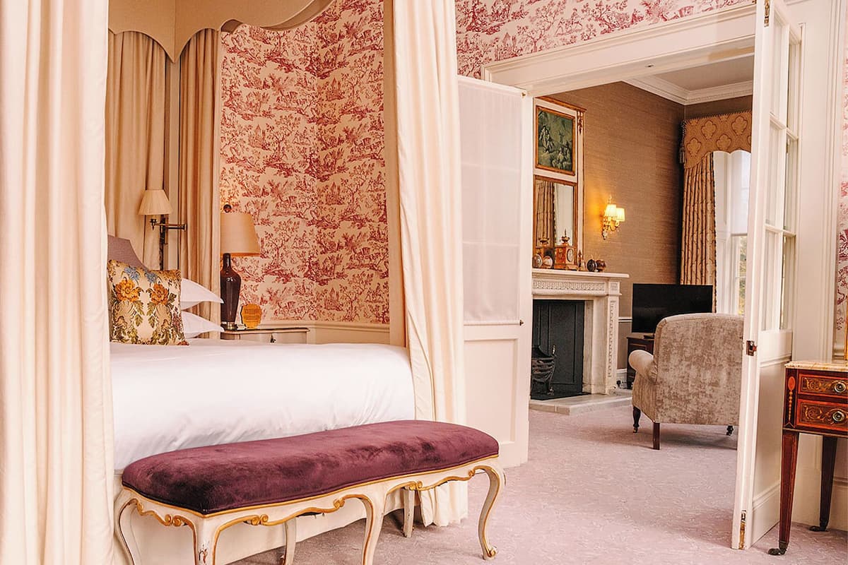 A bedroom with a four-poster bed, a bench, a chair, and a fireplace.