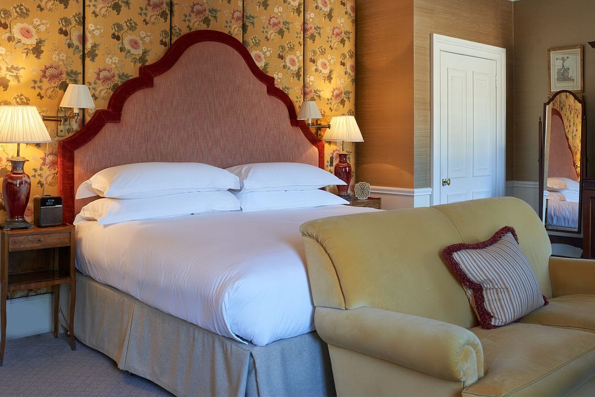 A bedroom with a large bed, two bedside lamps, a wooden headboard, a cream-colored armchair, and a floral wallpapered wall.