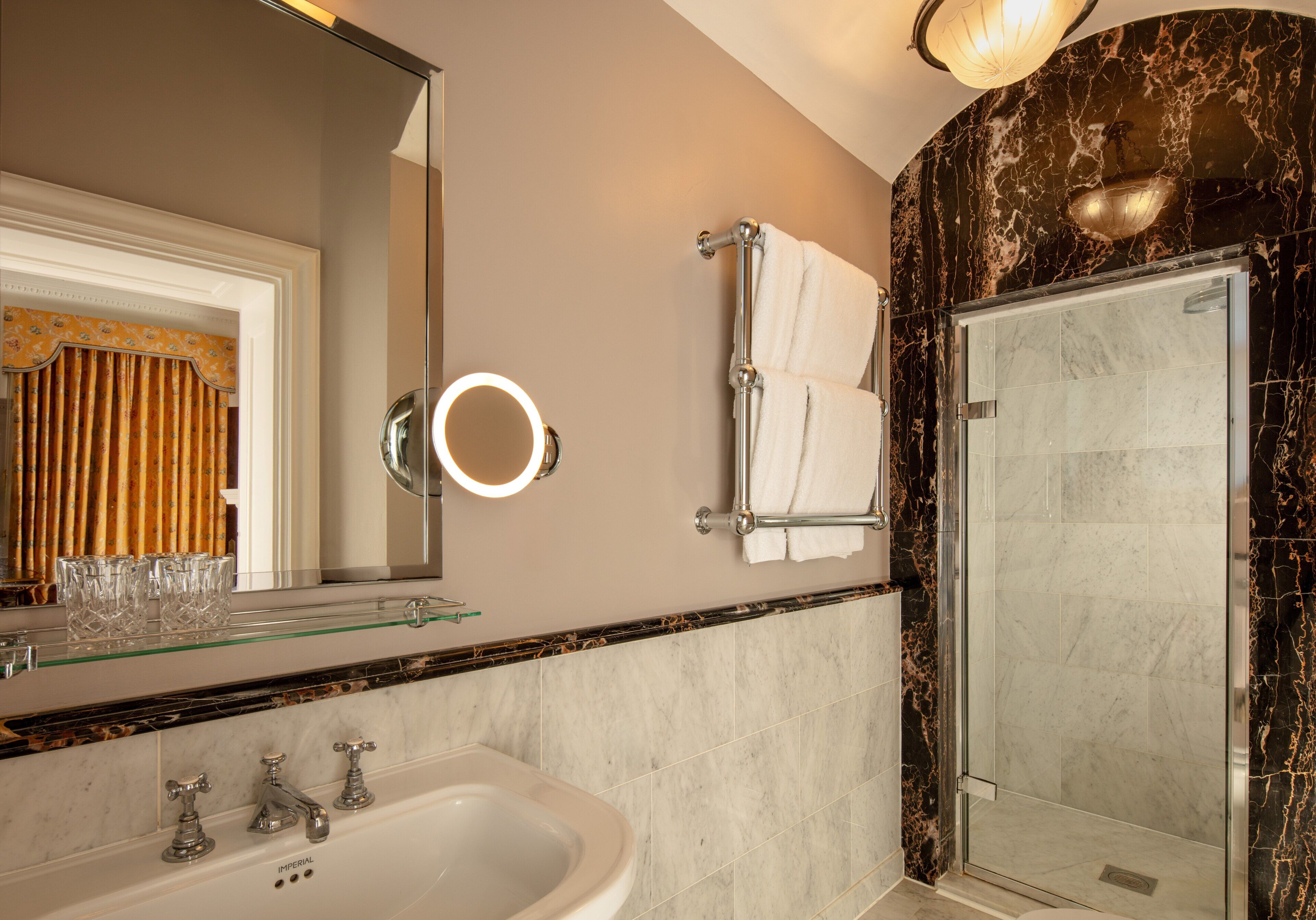 A bathroom with a glass shelf, a sink, a mirror with a light, a towel rack, and a shower area.