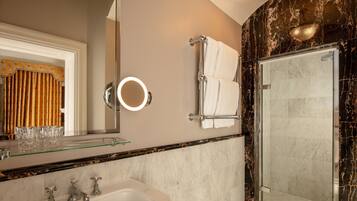 A bathroom with a glass shelf, a sink, a mirror with a light, a towel rack, and a shower area.