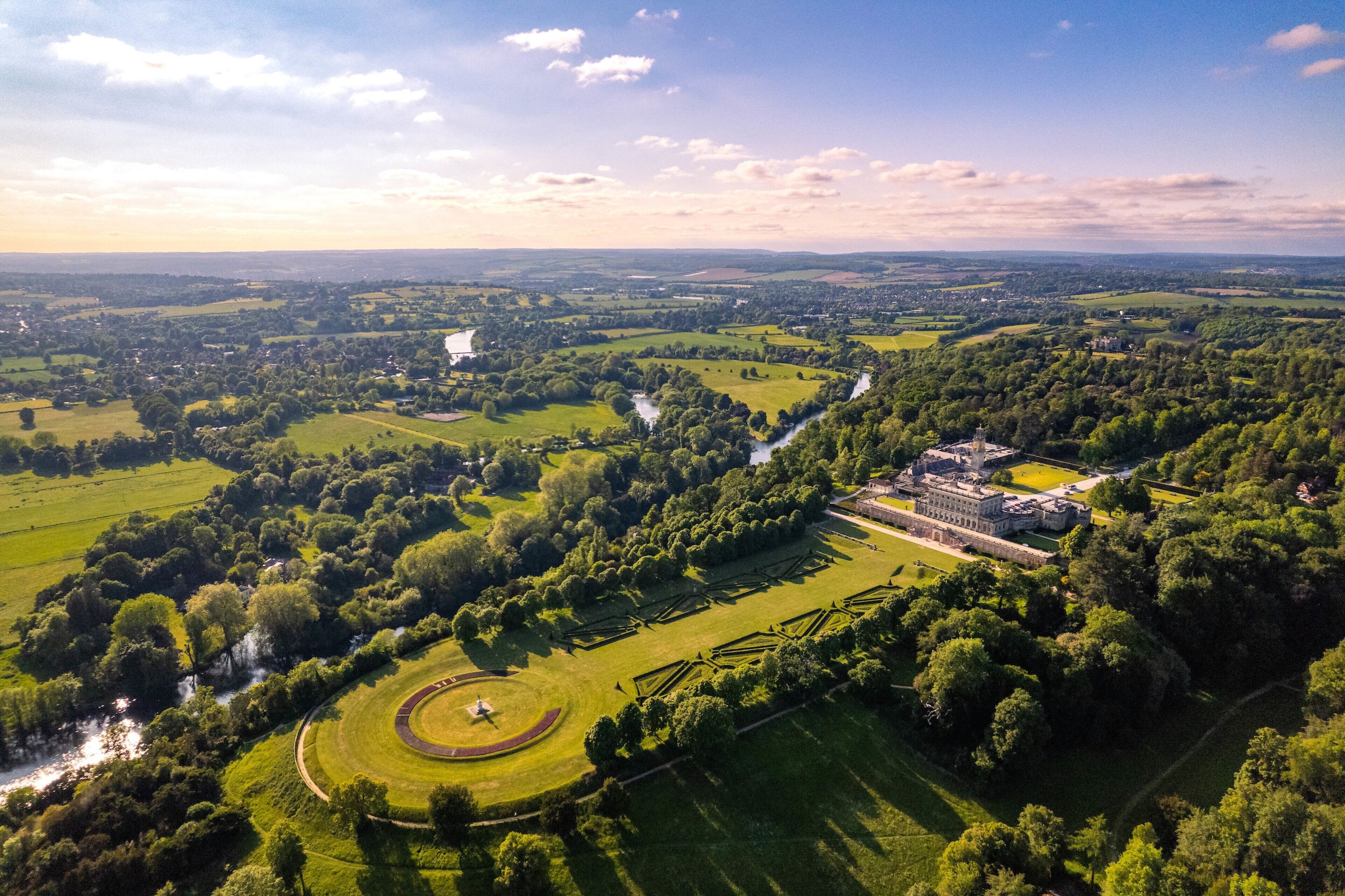 An aerial view of a historic estate with expansive green fields, a central circular lawn, and a large building with a formal garden.