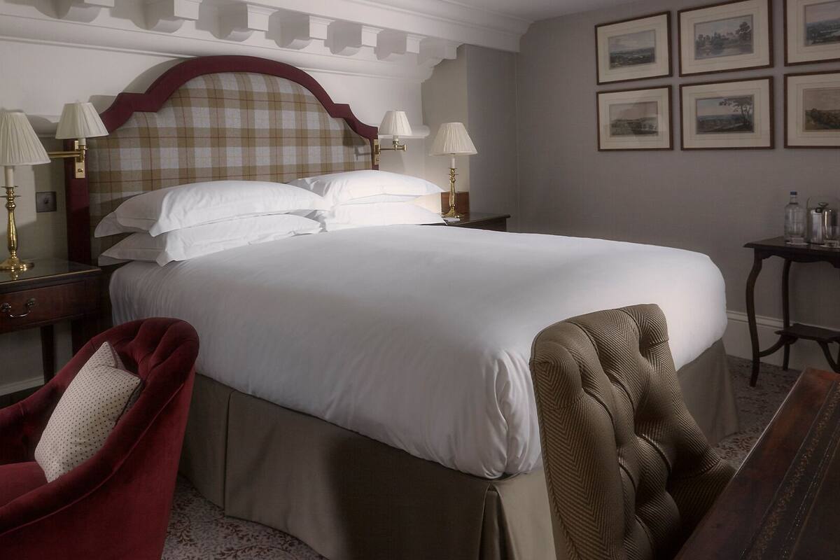 A neatly made bed with white linens, a red upholstered chair, a wooden nightstand with lamps, and a small table with bottles.