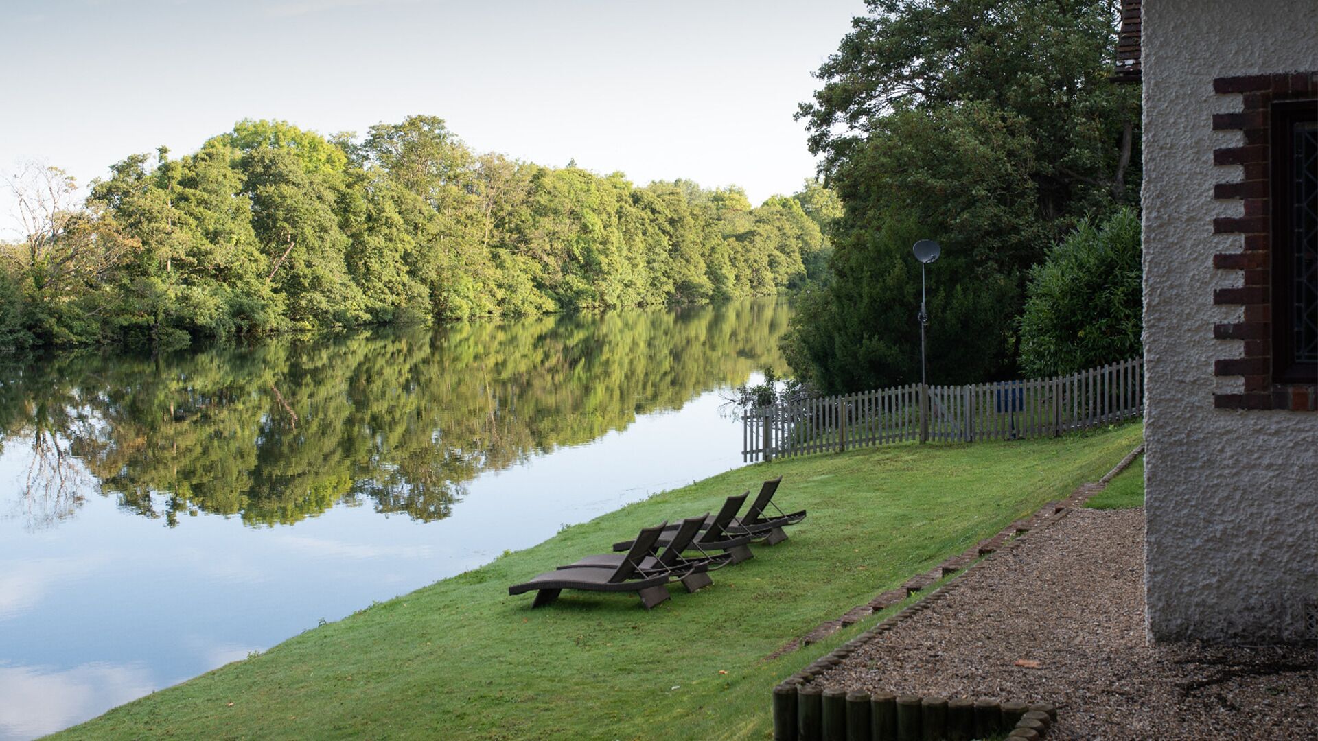 A lakeside view with a grassy area, wooden lounge chairs, and a building with a brick exterior.
