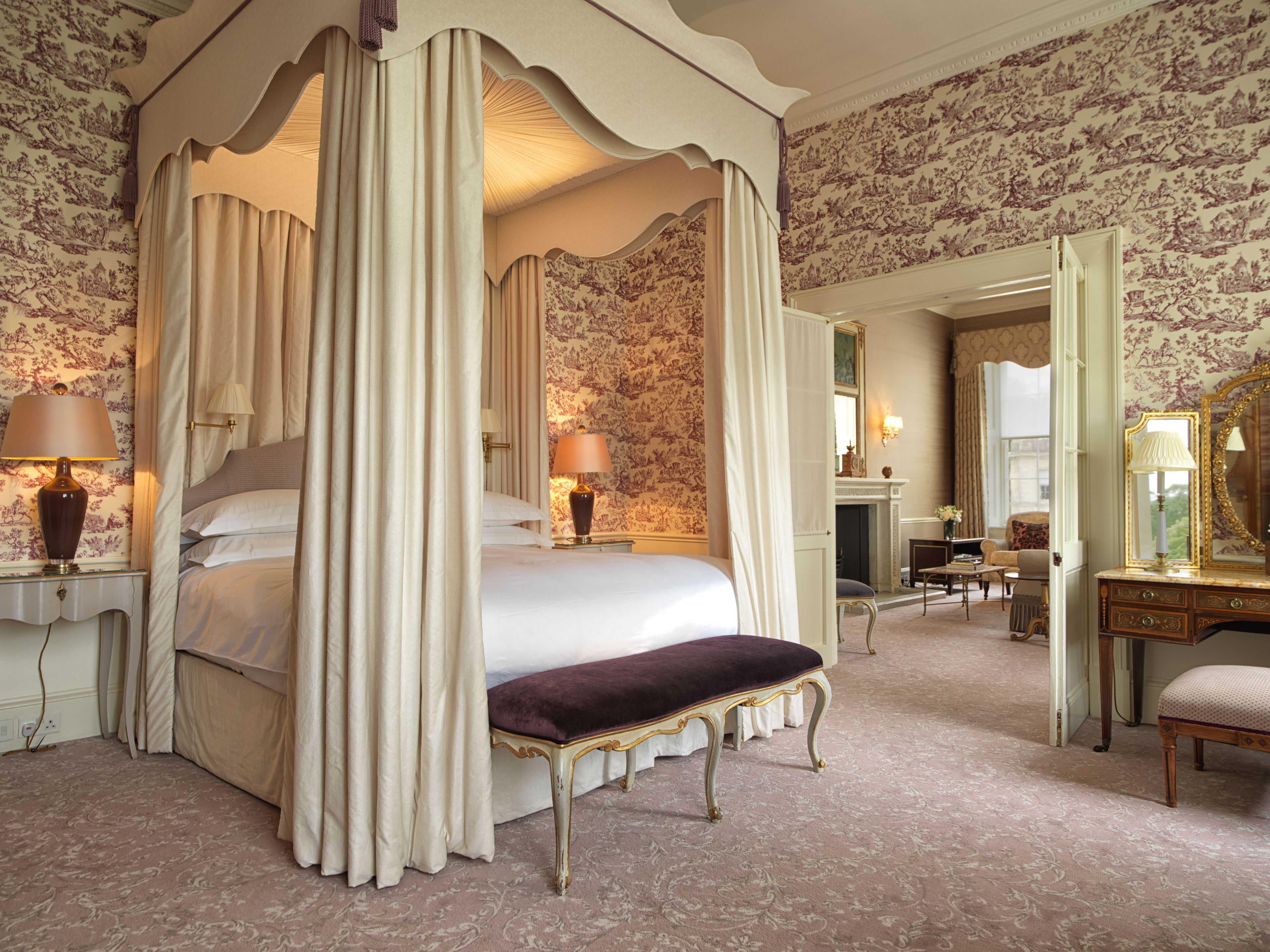 A four-poster canopy bed with a bench, a fireplace, and a mirror in a room with floral wallpaper.