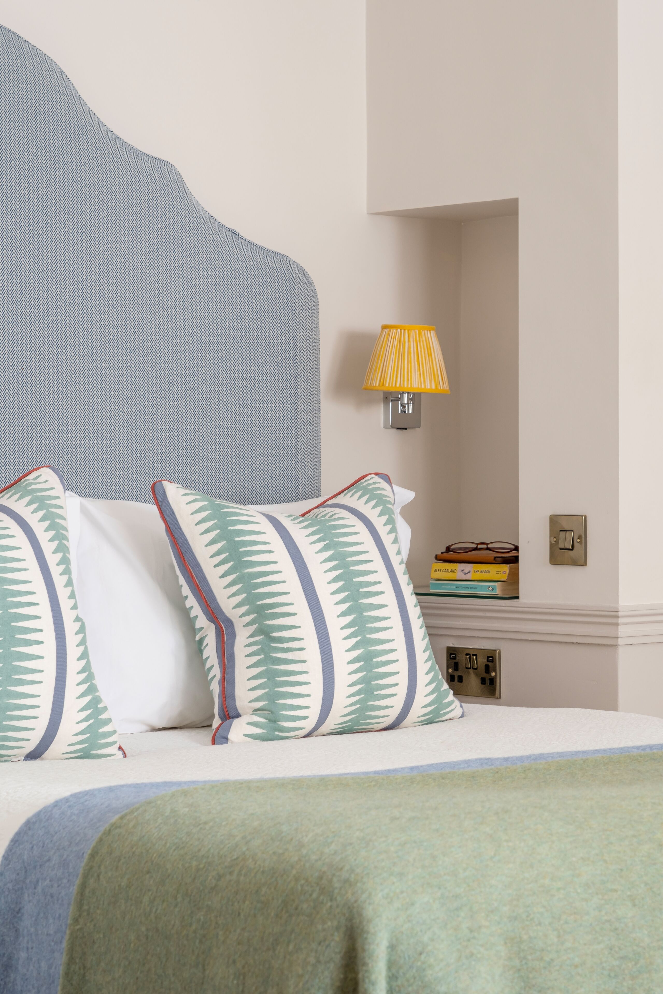 Een net opgemaakt bed met gestreepte kussens en een groene deken. Op het nachtkastje staat een bedlamp en een stapel boeken.