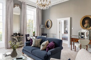 A living room with a blue sofa, a glass coffee table, and a chandelier. There is a mirror, a dresser, and a chair in the room.