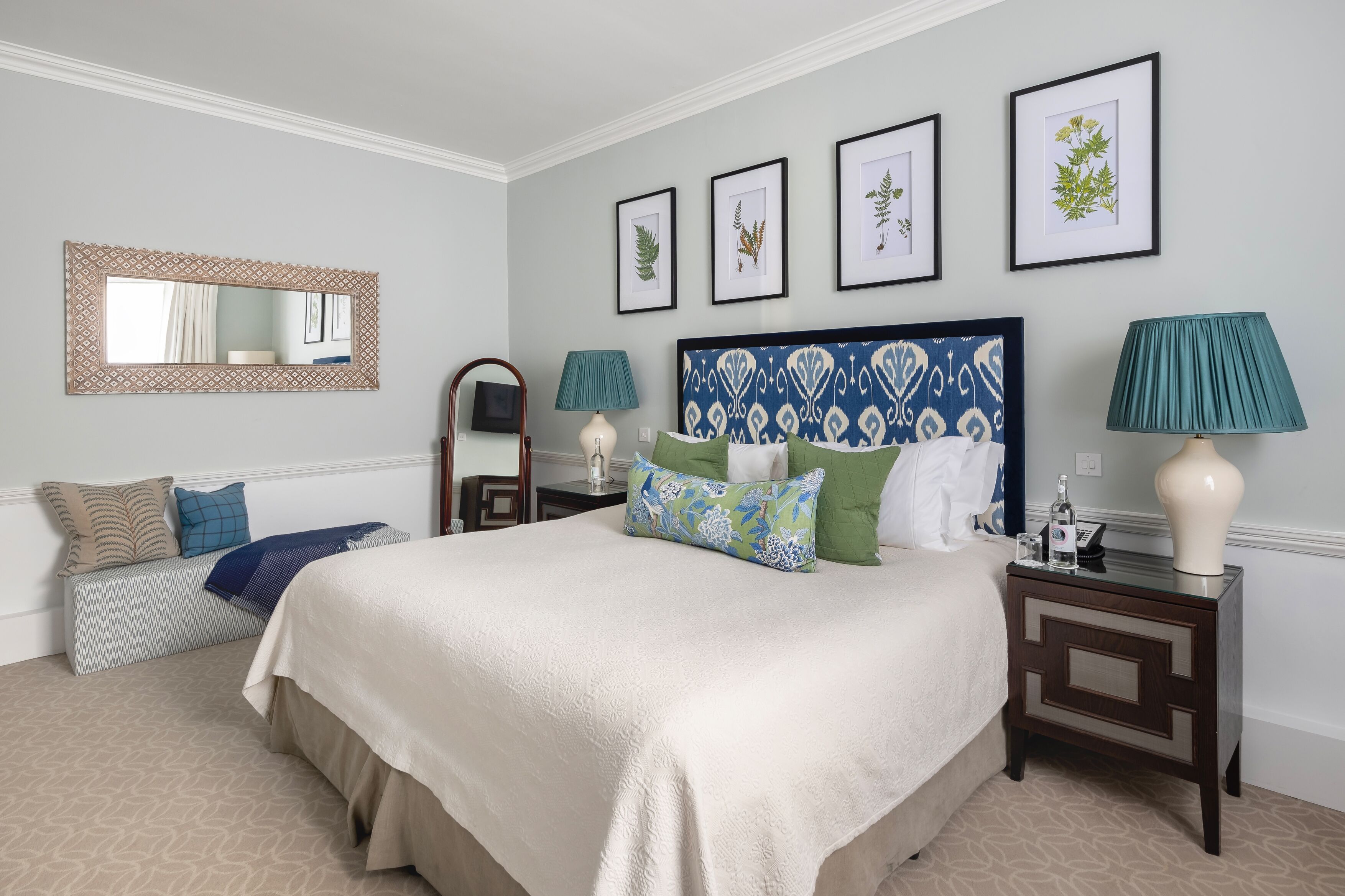 A neatly arranged bedroom with a large bed, a bedside table with lamps, a mirror, and framed botanical prints on the wall.