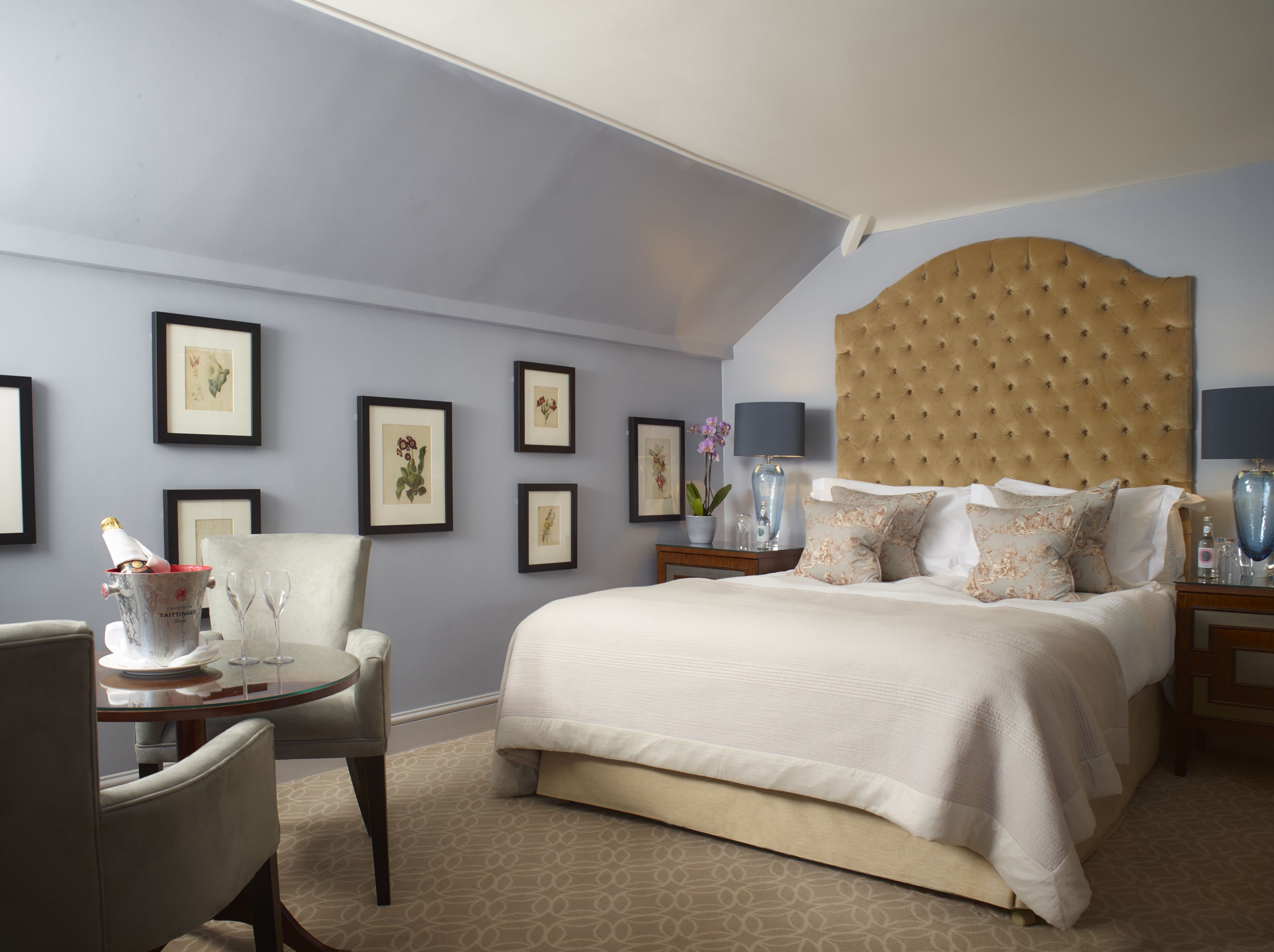 Habitación de hotel con una cama grande, una silla, una mesita con un florero y vasos, y cuadros en la pared.