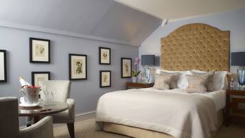 Habitación de hotel con una cama grande, una silla, una mesita con un florero y vasos, y cuadros en la pared.