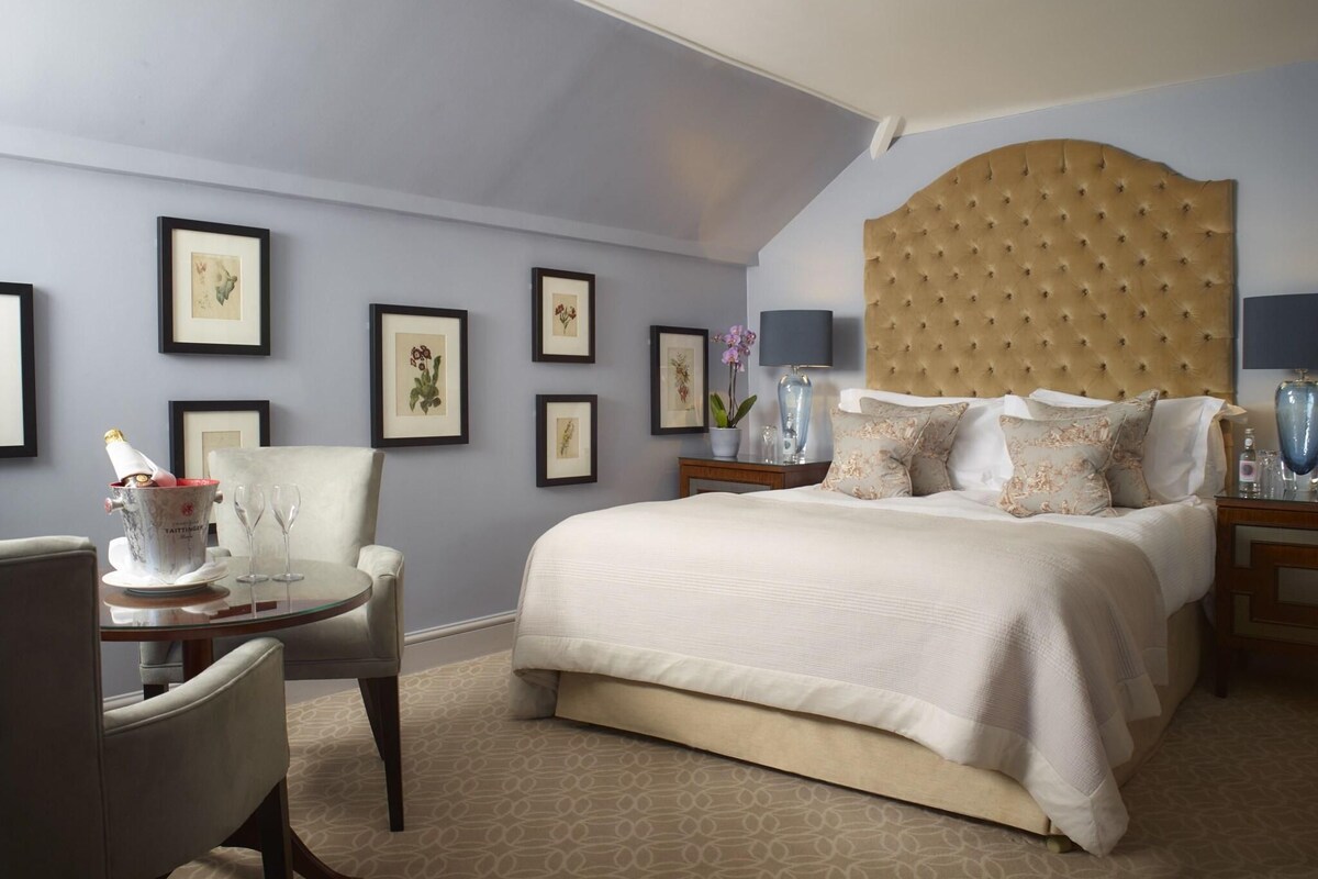 A hotel room with a large bed, a chair, a small table with a bottle and glasses, and framed pictures on the wall.