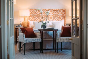 Habitación de hotel con una cama, dos sillones estampados, una mesita con una planta en maceta y una mesita de noche con una lámpara y una botella.