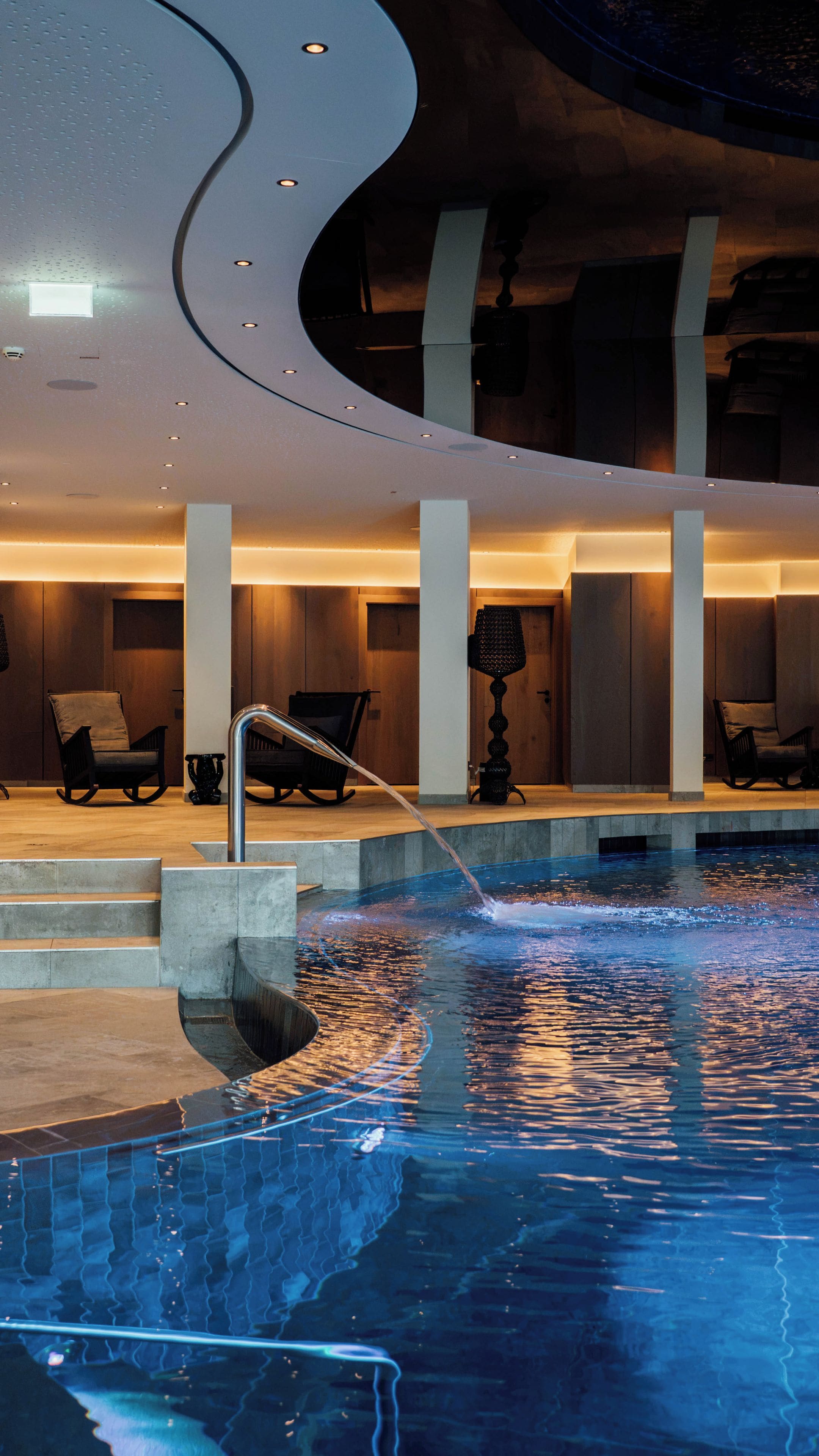 Un área de piscina interior moderna con superficie de agua cristalina, escalones para entrar y zonas de estar con sillas.