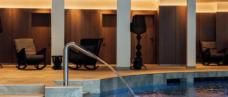 Un área de piscina interior moderna con superficie de agua cristalina, escalones para entrar y zonas de estar con sillas.