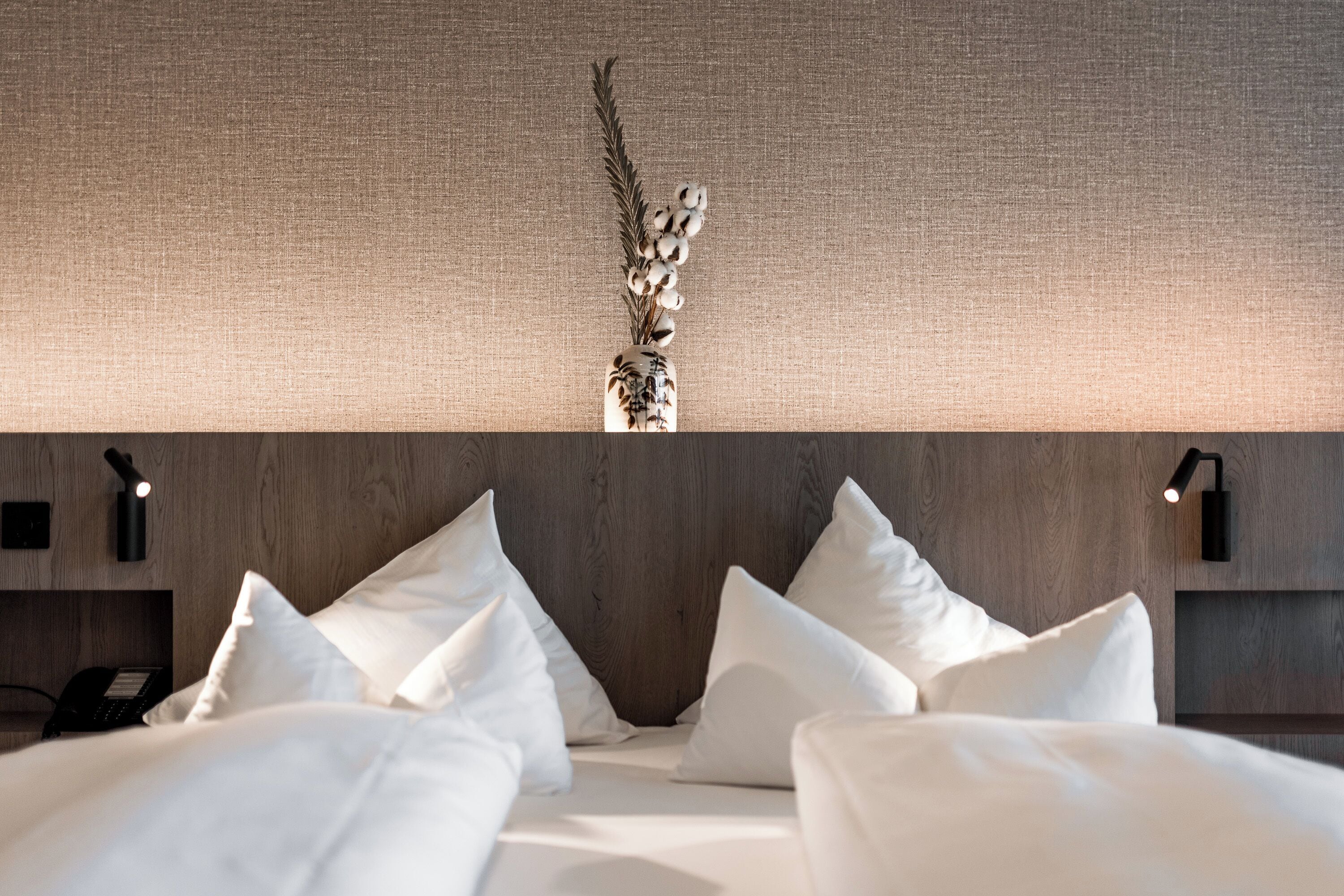 Una habitación de hotel con una cama, almohadas, un florero con una planta y luces fijadas en la pared.