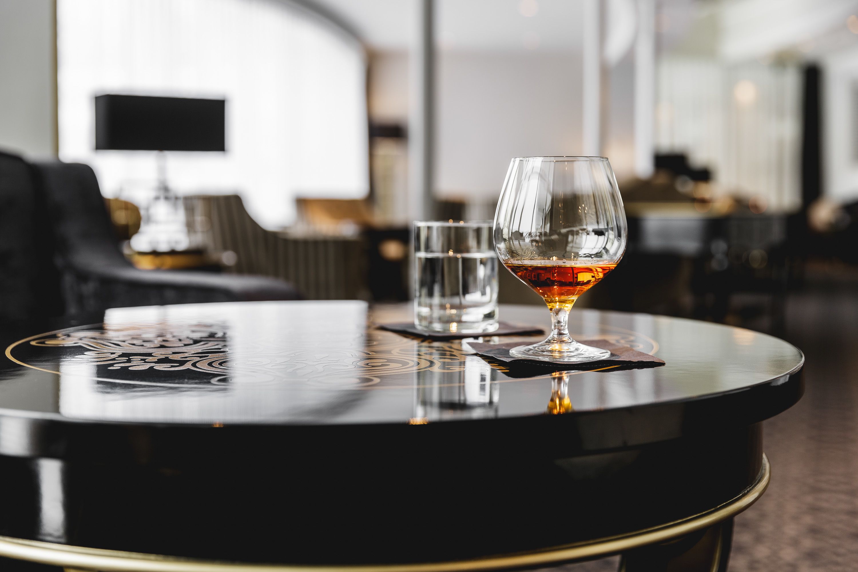 A round table with a glass of amber liquid and a clear glass of water, a black lamp, and a patterned tablecloth.