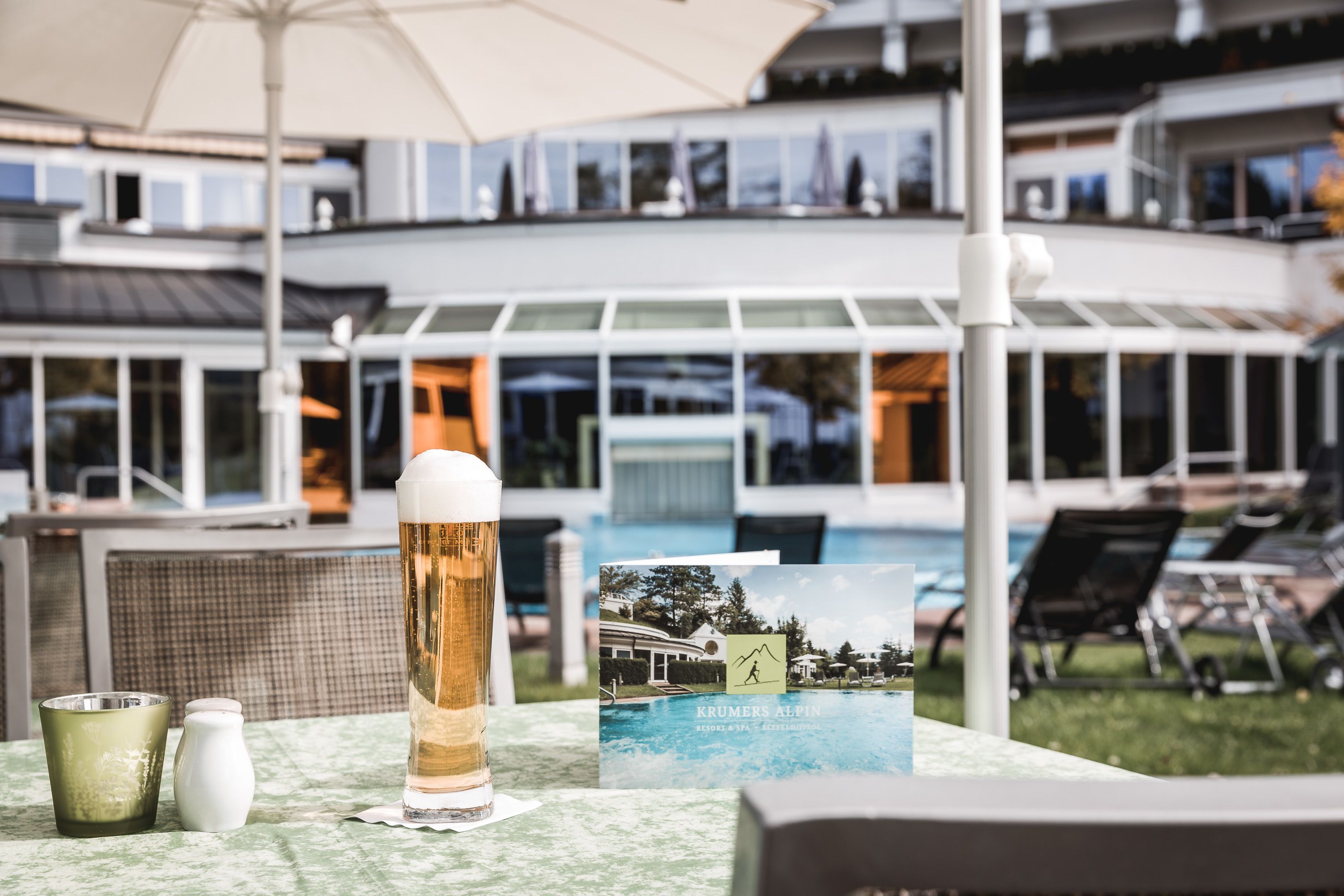 A table with a beer glass, a salt shaker, and a postcard at a poolside area with lounge chairs and a pool in the background.