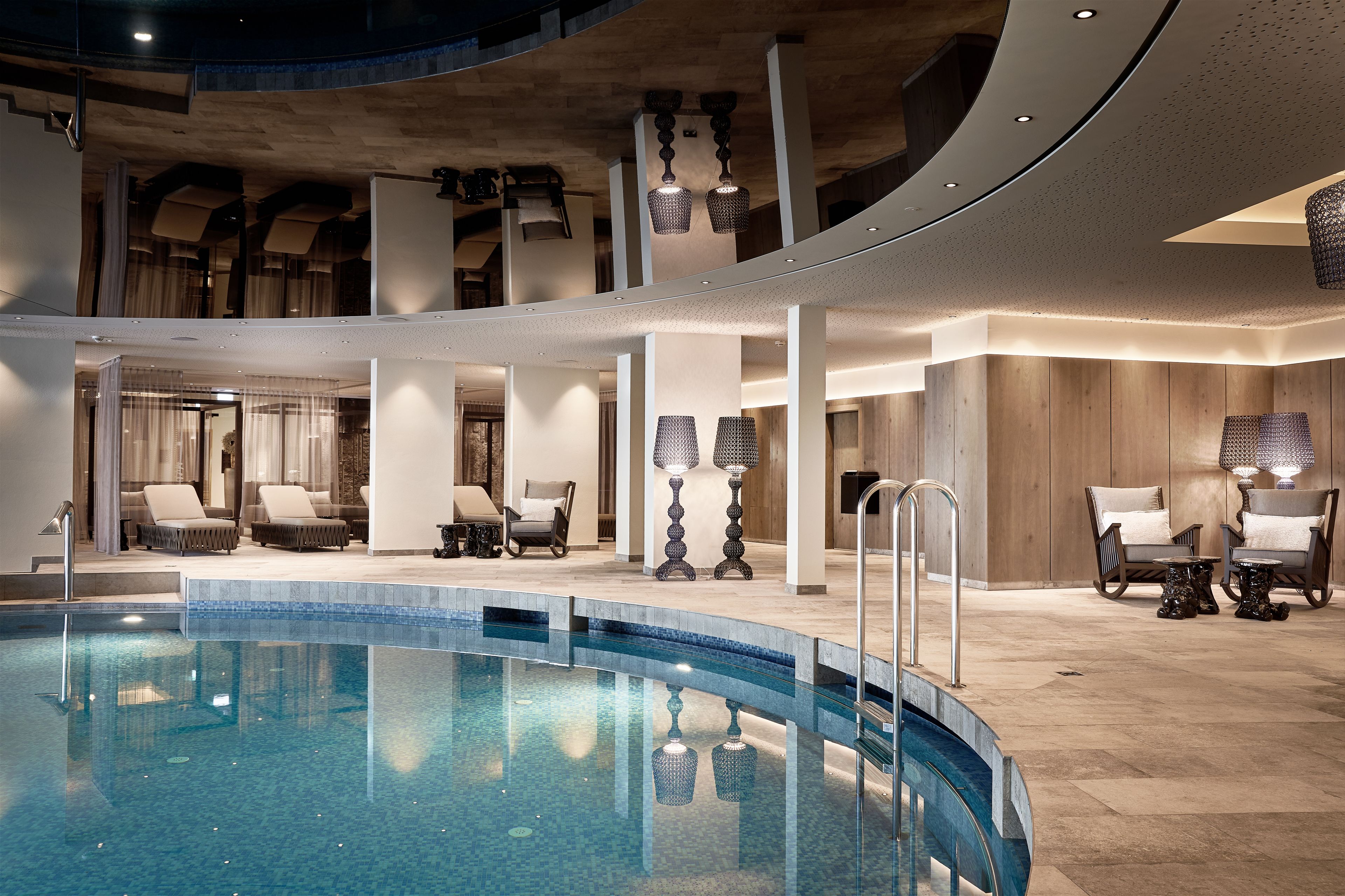 Une piscine intérieure moderne, dotée de chaises longues et de tables, et entourée de panneaux de bois et d’un éclairage contemporain.