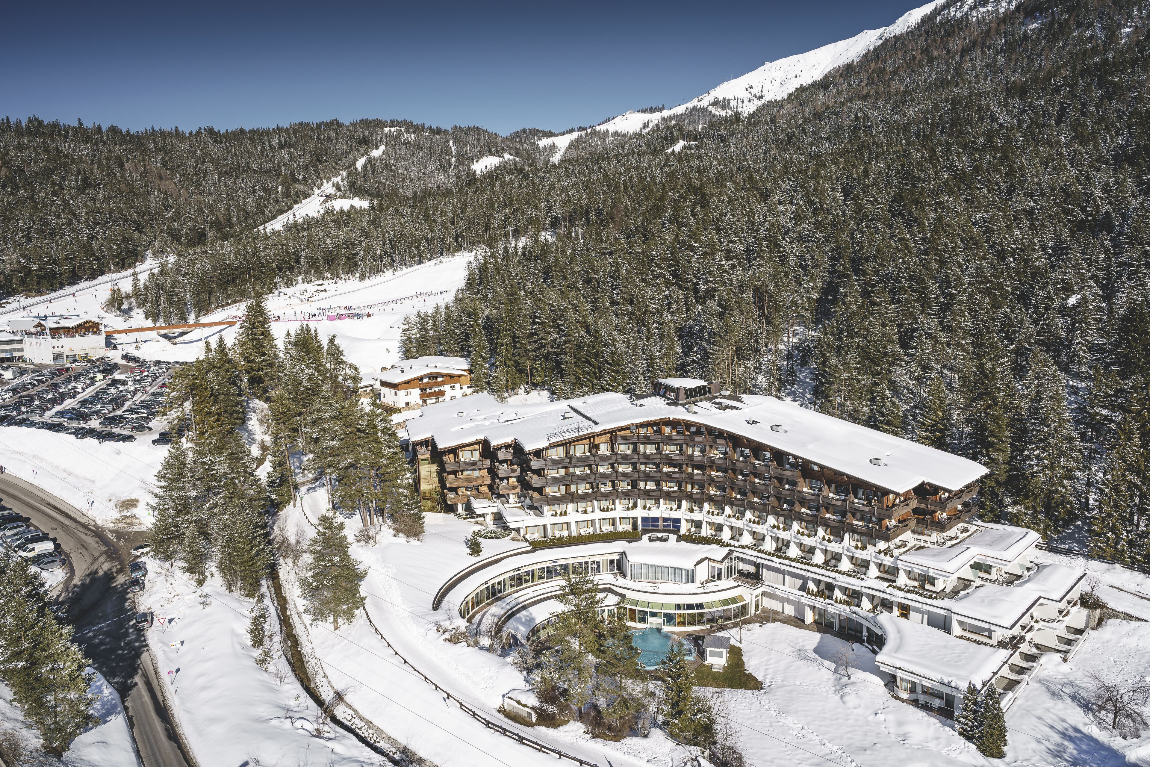 A large, multi-story hotel with a curved facade, surrounded by snow-covered mountains and trees.