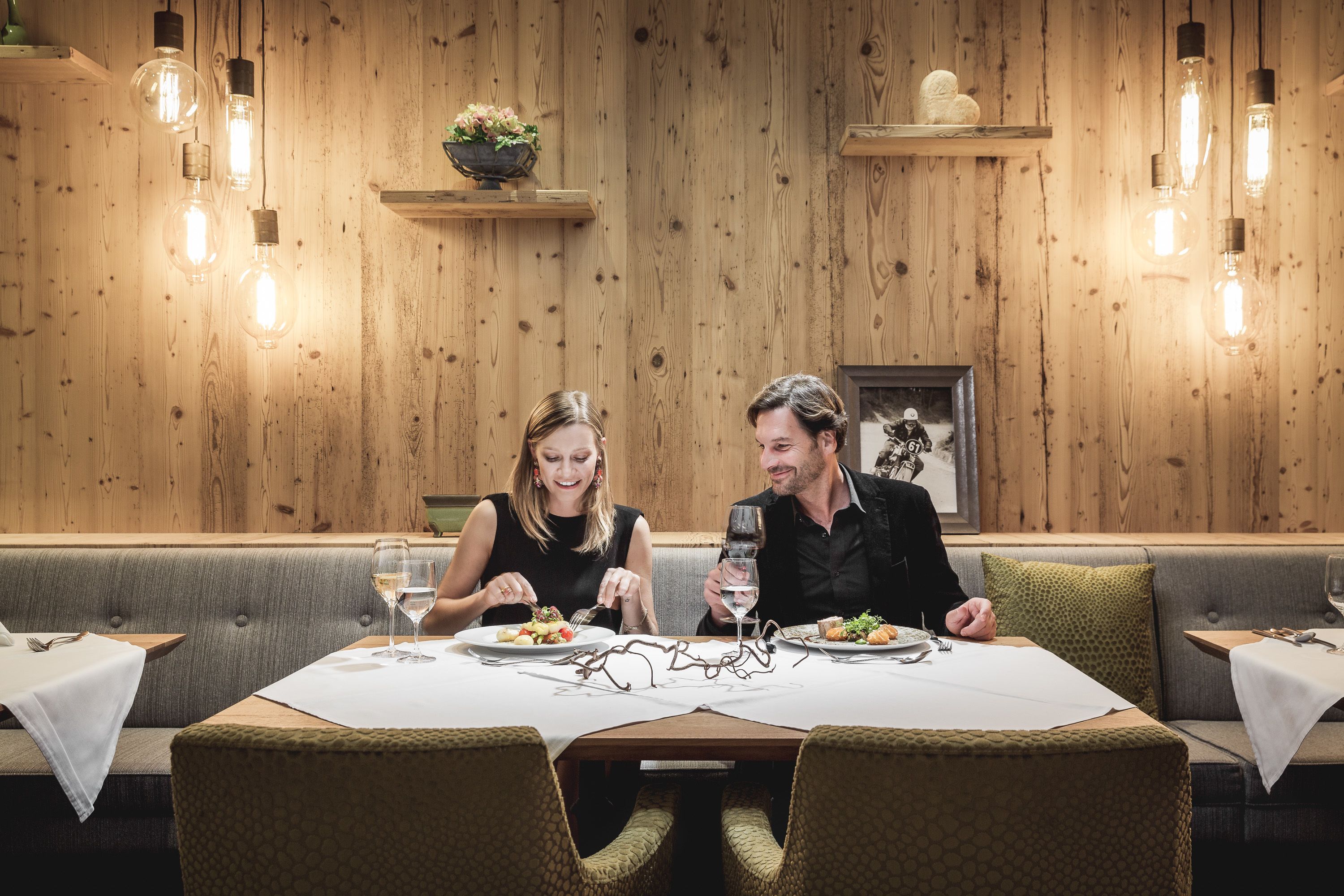 Una pareja cenando en un restaurante con interior de madera, iluminación colgante y mantel blanco.