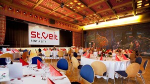 Een balzaal met ronde tafels gedekt voor een formeel evenement, een podium met een spandoek en een muurschildering.