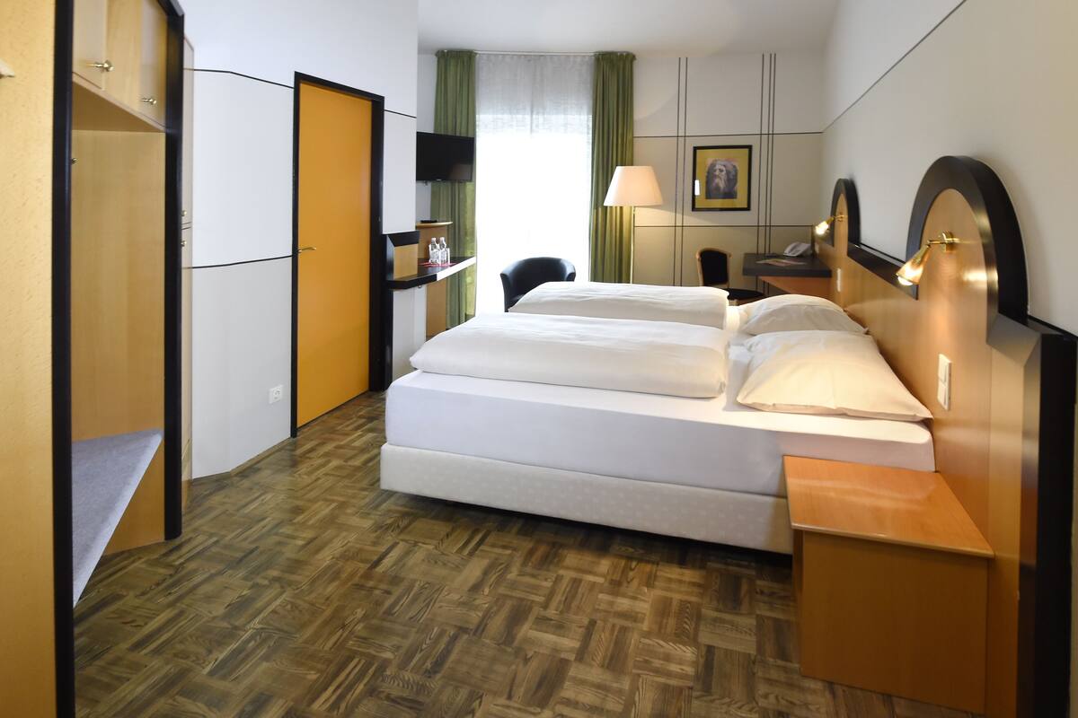 Una habitación de hotel con cama, escritorio, silla, armario y televisor en la pared.