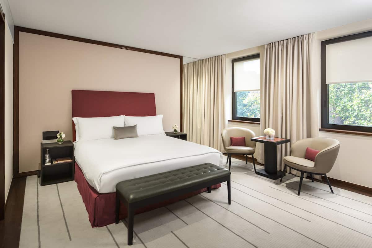 Une chambre d’hôtel avec un grand lit, une table de chevet, une chaise, une petite table et une fenêtre avec des rideaux.