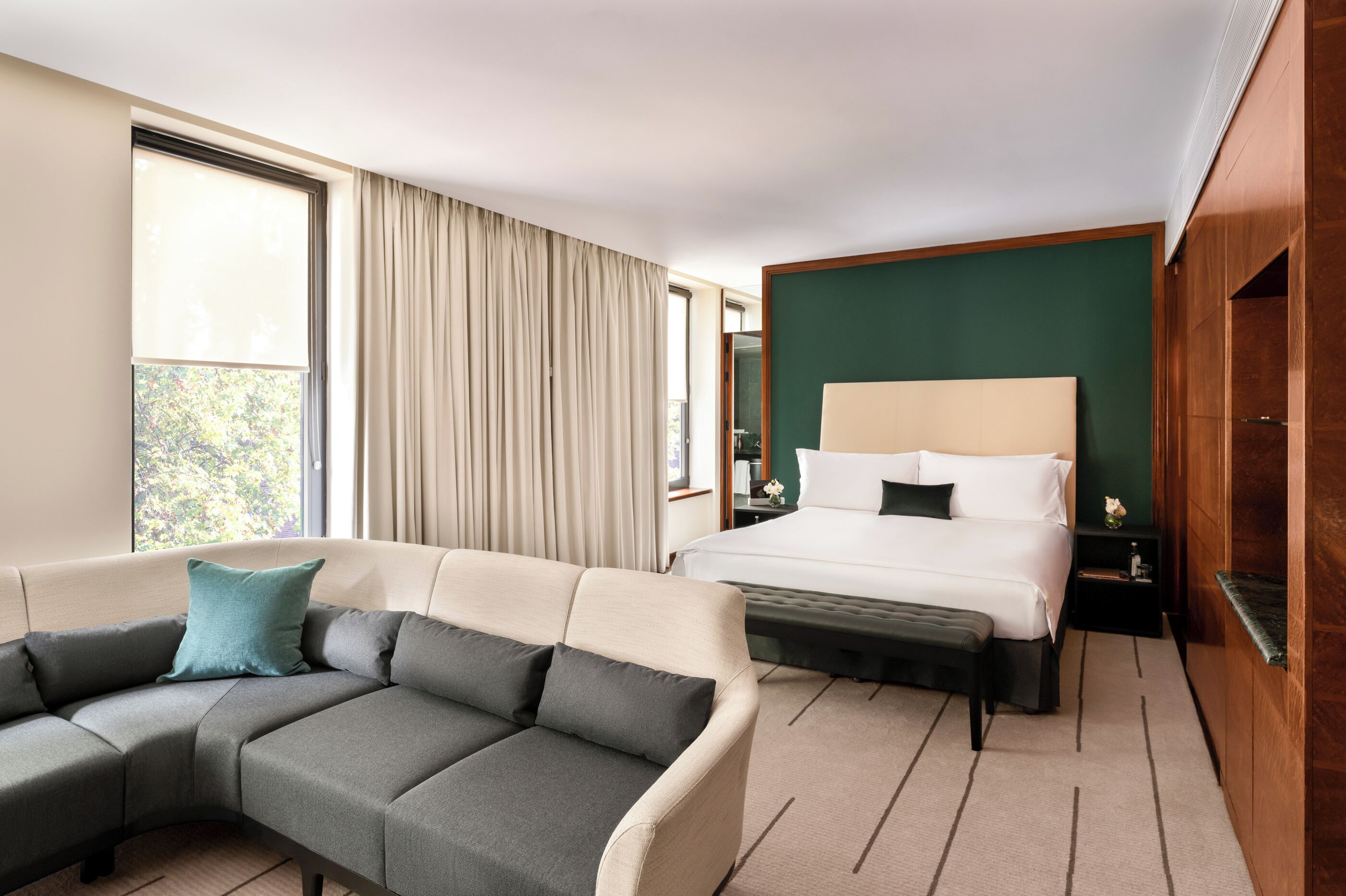 A hotel room with a sectional sofa, a bed with a headboard, a nightstand, and a view through a window.