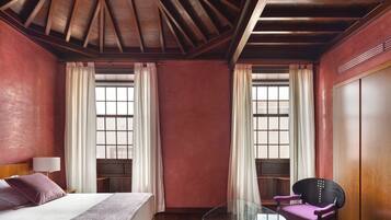 Una habitación de hotel con techo de madera, una cama con edredón blanco, una mesa de centro de vidrio, una silla morada y un armario.