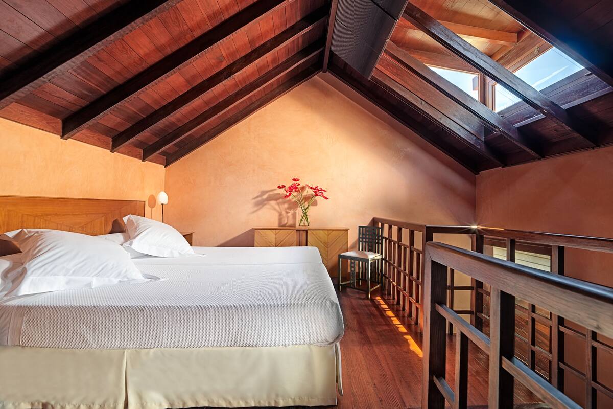 Un dormitorio con techo de madera, una cama, una mesita de noche, un jarrón con flores y vistas al cielo a través de tragaluces.