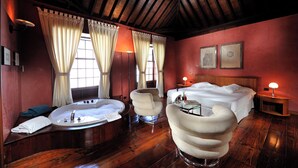 Una habitación con una cama, una bañera redonda, dos sillones, una mesita de noche con una lámpara y una ventana con cortinas.