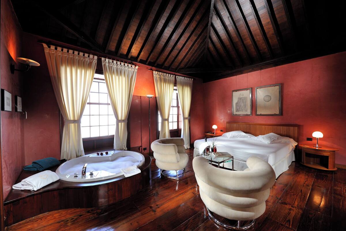 Una habitación con una cama, una bañera redonda, dos sillones, una mesita de noche con una lámpara y una ventana con cortinas.
