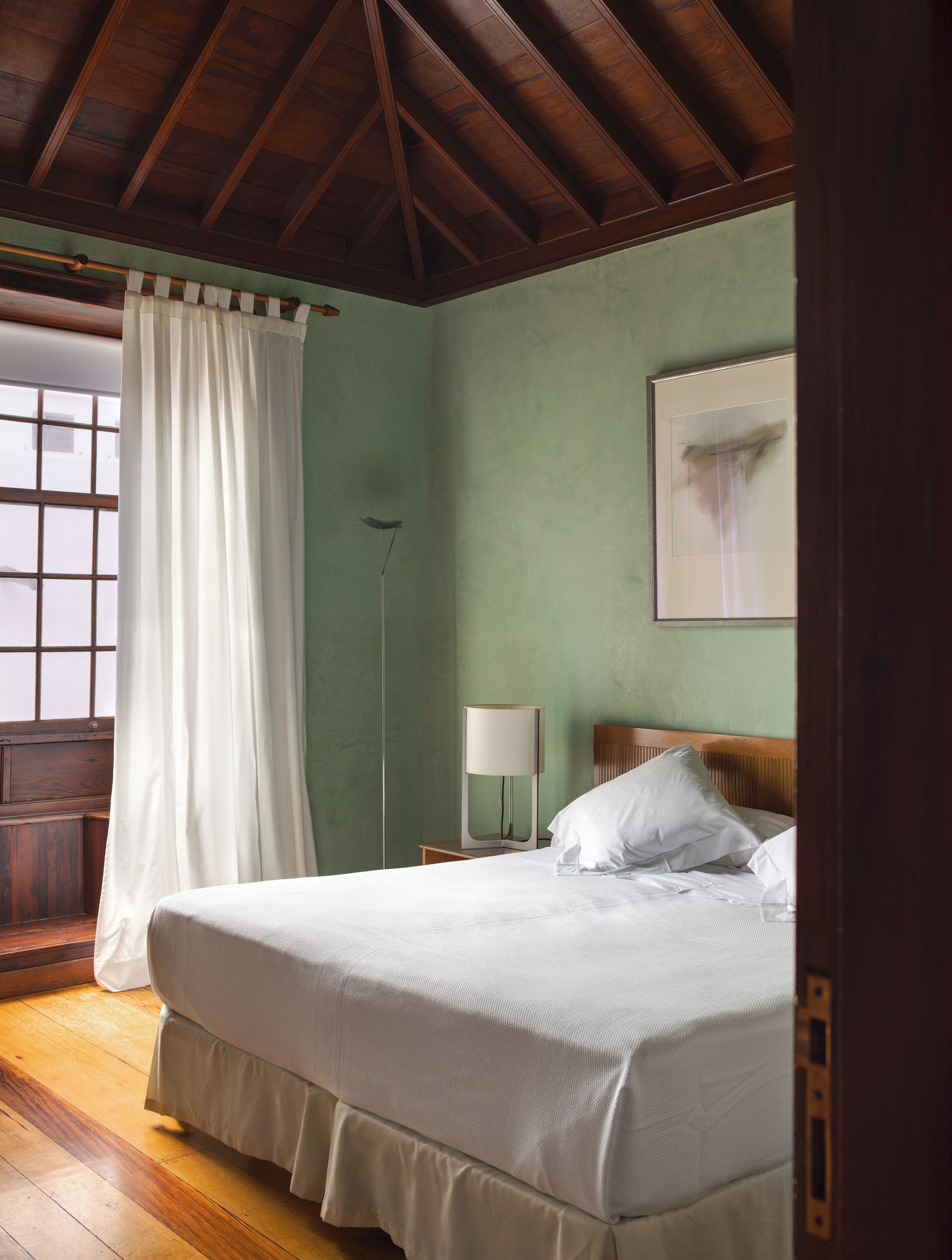 A bedroom with a wooden ceiling, a bed with white bedding, a bedside table with a lamp, and a window with curtains.
