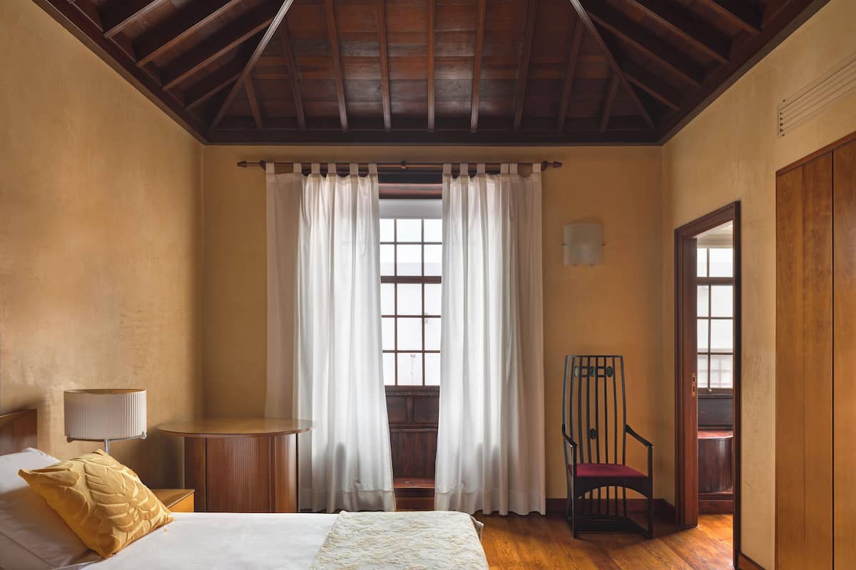 Un dormitorio con techo de madera, una cama con colcha blanca, una mesita de noche con lámpara y una silla.