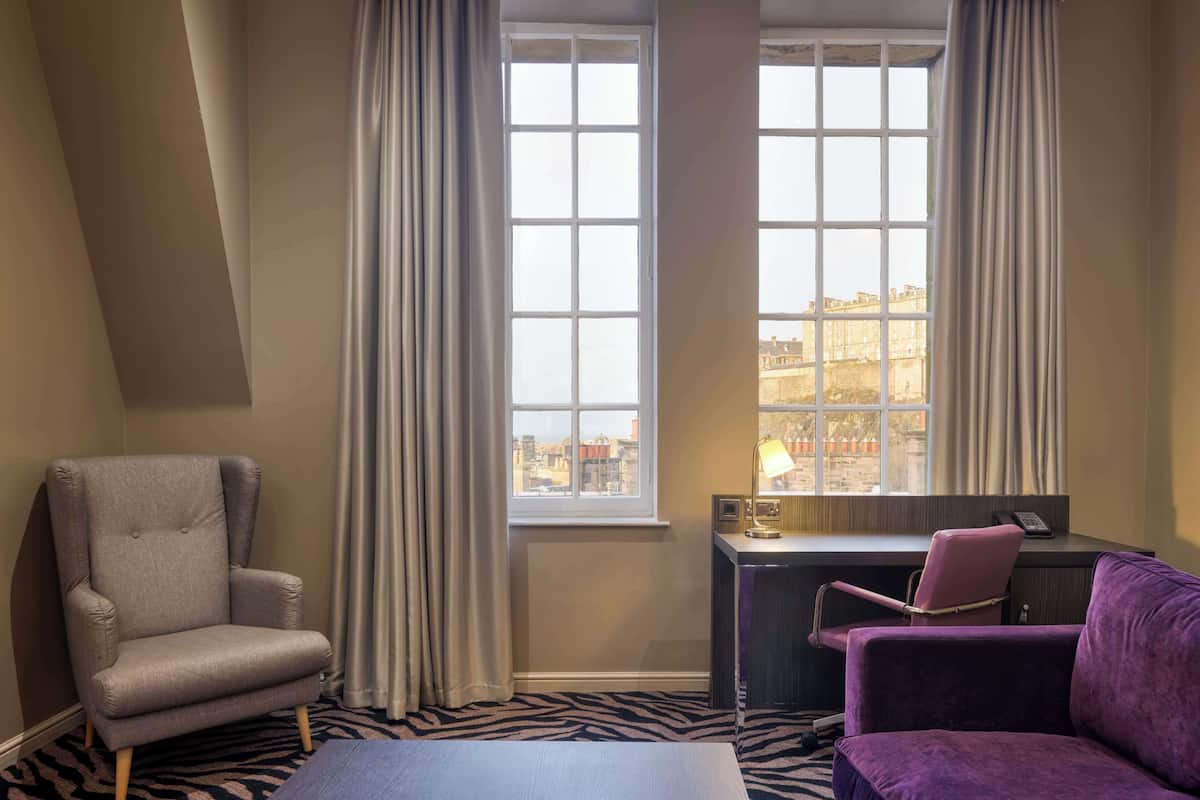 A hotel room with a grey armchair, a purple sofa, a desk with a lamp, and a view of buildings through large windows.