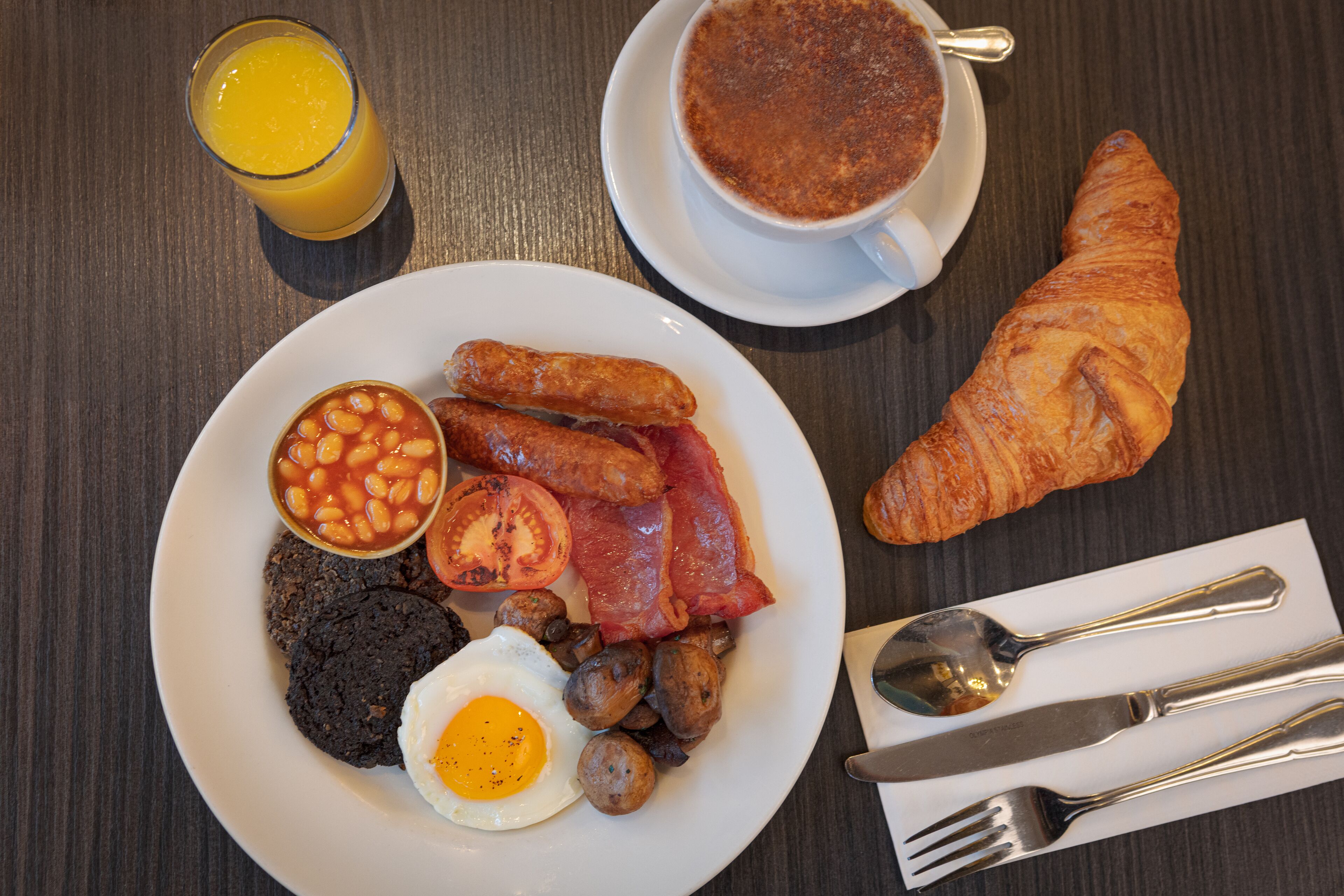 Une assiette de petit-déjeuner comprenant des œufs, des saucisses, des haricots, du bacon, des champignons et des toasts, ainsi qu’un croissant et une tasse de café.